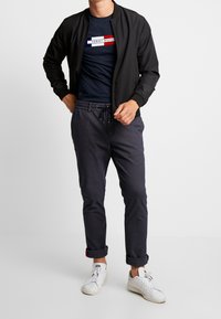 Muž v černé bomber bundě, námořnickém tričku s logem Tommy Hilfiger, tmavě šedých zúžených kalhotách a bílých teniskách, stojí na bílém pozadí.