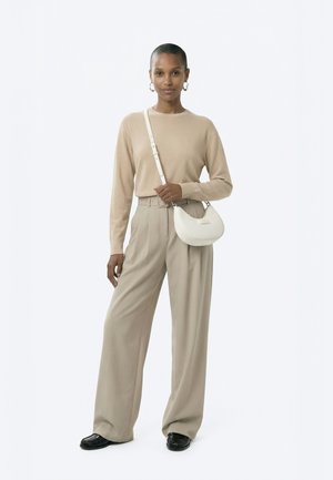 Žena u bež džemperu i širokim hlačama, nosi malu bijelu torbicu preko ramena, stoji ispred jednobojne bijele pozadine.