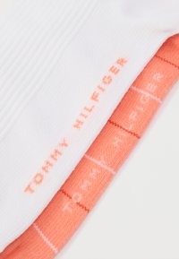 Δύο ζευγάρια κάλτσες, ένα λευκό με ριγέ υφή και ένα κοραλί με σχέδιο πλέγματος, και τα δύο με το λογότυπο «TOMMY HILFIGER».