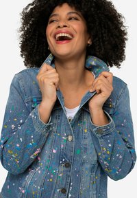 Veste en denim avec un délavage bleu clair, éclaboussures pulvérisées en rose, vert et jaune. Fermeture à boutons avec deux poches.