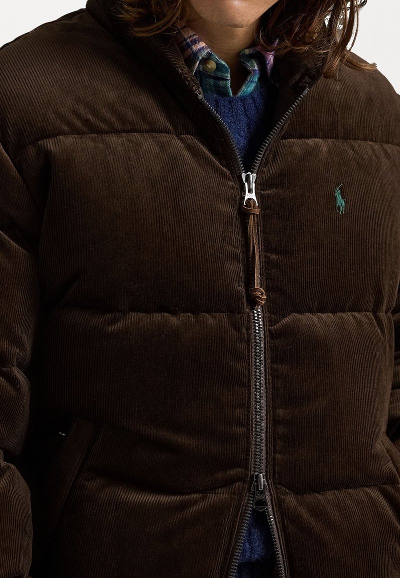 Veste matelassée en velours côtelé marron avec fermeture éclair, arborant un logo teal, un tissu texturé et une capuche à cordon de serrage. Superposée sur un pull bleu.