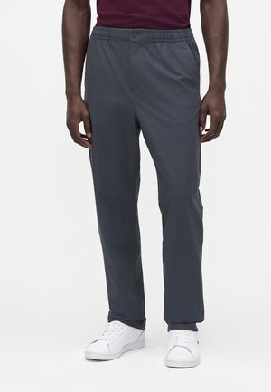 Uomo che indossa pantaloni grigio scuro con vita elastica e sneakers bianche, in piedi davanti a uno sfondo chiaro e uniforme.