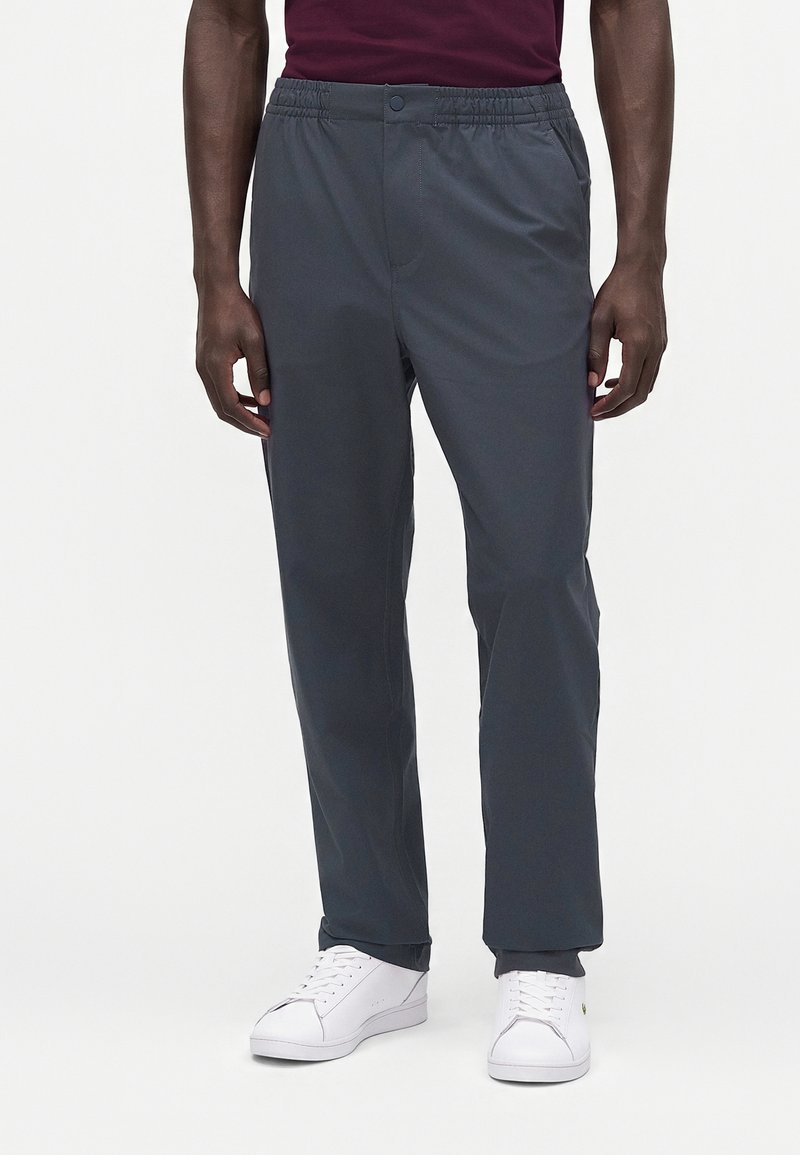 Homme portant un pantalon gris foncé à taille élastique et des baskets blanches, debout devant un fond clair uni.