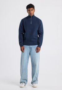 Navy zip-up sweater with high collar, ribbed hem, and cuffs; paired with light blue wide-leg jeans and white sneakers.