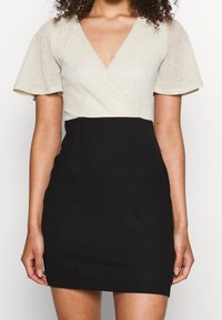 Robe courte avec un haut brillant couleur or clair et une jupe noire. Présente un col en V et des manches courtes évasées. Texture lisse sur l'ensemble.