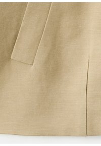 Tissu beige texturé avec un gros plan sur une couture de poche et une fente verticale sur un vêtement.