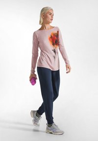 Langärmliges pinkes Oberteil mit Blumenmuster, kombiniert mit dunkelblauen Jogginghosen. Graue Sneakers mit grünen und lila Akzenten. Hält eine lila Wasserflasche.