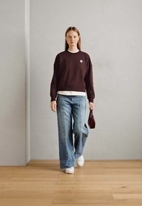 Sudadera burdeos con un pequeño logo, camisa blanca con capas, vaqueros de pierna ancha azul claro, zapatillas blancas y un bolso pequeño burdeos.