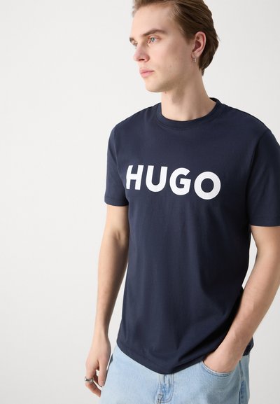 Нави синьо памучно тениска с голямо бяло лого "HUGO" отпред. Къси ръкави, кръгло деколте и свободен дизайн.