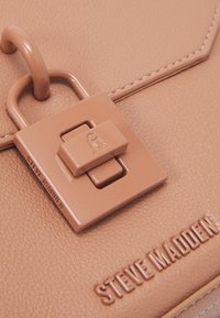 Růžová kabelka vyrobená z texturované umělé kůže. Má uzávěr na zámek, výrazné logo a hladký, lesklý povrch.