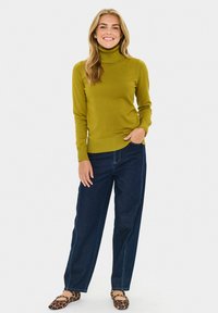 Olivno zelena turtleneck pulover s rebrastim robom, v paru s temno modrimi širokimi hlačami in balerinkami z živalskim potiskom, roke v žepih.