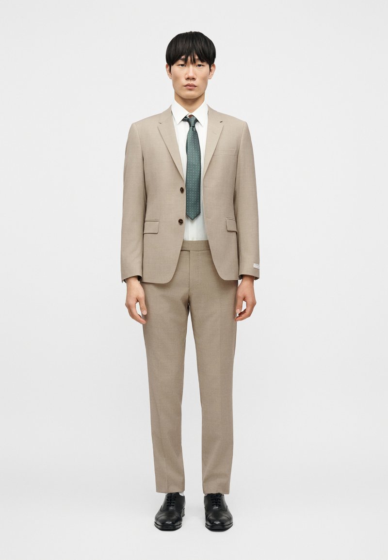 Μπεζ κοστούμι με εφαρμοστή σακάκι  και παντελόνι ίσιας γραμμής, συνδυασμένο με λευκό πουκάμισο και πράσινη γραβάτα με μικρό μοτίβο. Μαύρα παπούτσια.