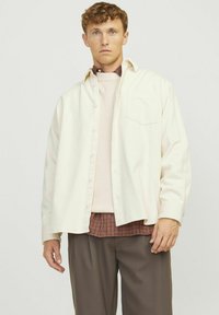 Camicia di corduroy crema, con bottoni e una tasca sul petto; indossata sopra un maglione rosa chiaro e una camicia a quadri; abbinata a pantaloni marroni.