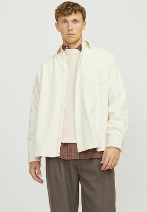 Cream corduroy shirt, button-up, with a chest pocket; layered over a light pink sweater and checked shirt; paired with brown trousers.