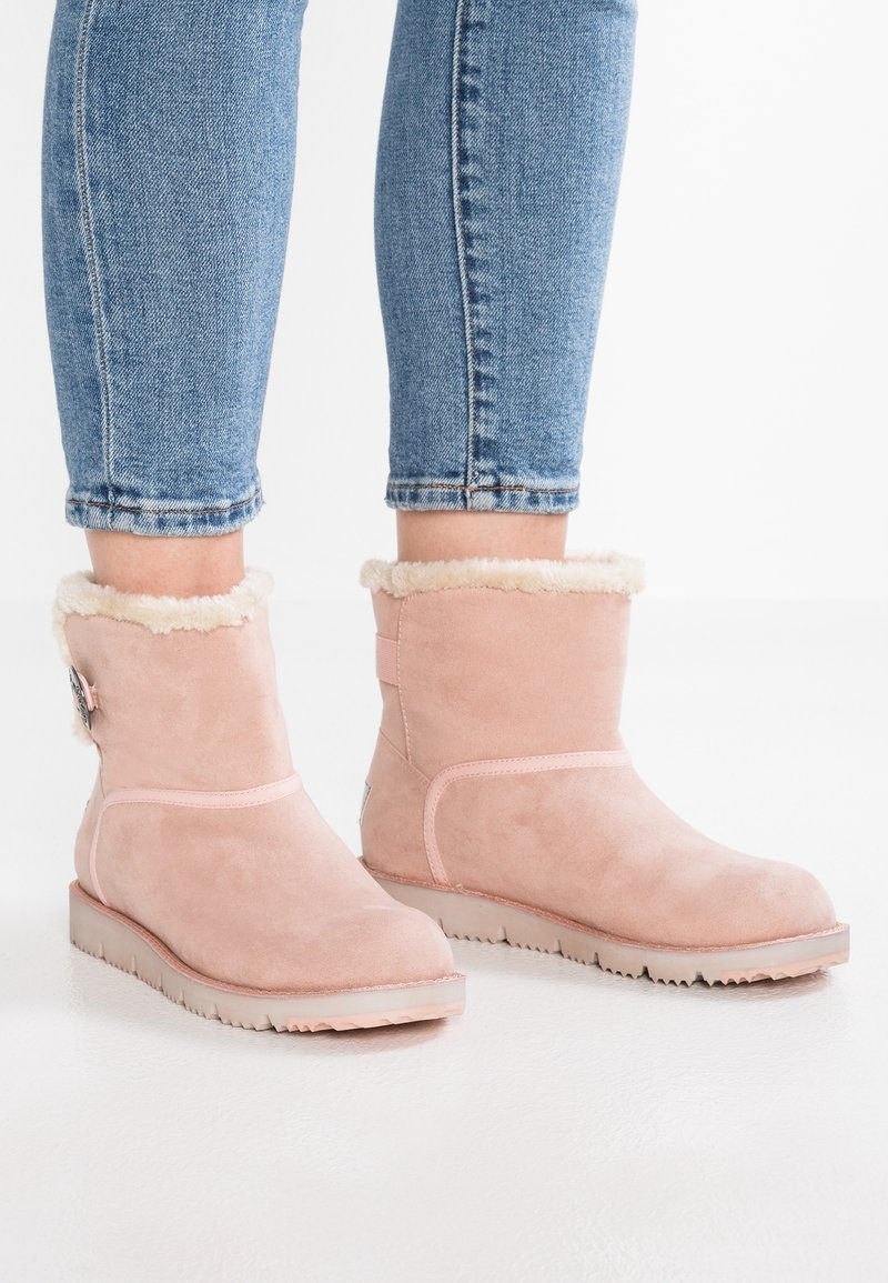 Bottines en daim rose clair avec doublure en fausse fourrure beige, portées avec un jean denim bleu retroussé sur un sol blanc.
