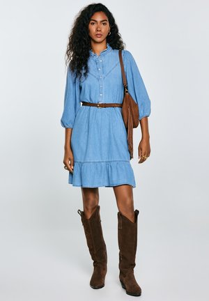 Light blue denim dress with a ruffled hem, button-down front, and gathered sleeves, paired with brown suede knee-high boots and a brown bag.