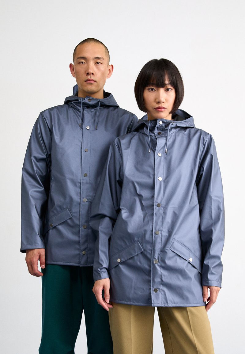 Des vestes imperméables grises avec capuches, boutons-pression et poches avant, portées par deux modèles sur un fond uni.