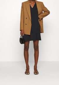 Blazer marrón de doble botonadura sobre un vestido negro. Un bolso negro y sandalias de estampado de leopardo complementan el atuendo. Textura suave y corte ajustado.