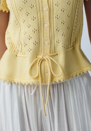 Gul strikket ærmeløs cardigan med knapper foran, hulmønster, snøre i taljen og bølget kant over en hvid plisseret nederdel.