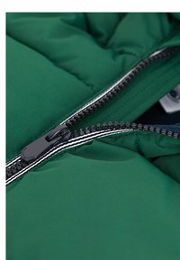 Grønn polstret jakke med svart glidelås og kontrasterende hvit stripe. Myk tekstur med quiltet design og indre stoff synlig nært glidelåsen.