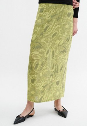 Lang, groene rok met abstracte gele en donkergroene patronen; gemaakt van gestructureerde stof. Draagt met zwarte, puntige ballerina's.
