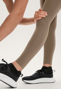Brązowe legginsy sportowe o gładkiej, matowej teksturze, w połączeniu z czarnymi sneakersami z siateczkowymi akcentami i miękką białą podeszwą.