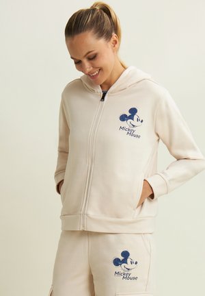 Femme portant un sweat à capuche beige zippé et un pantalon assorti avec des imprimés bleus de Mickey Mouse, les mains dans les poches, souriante et regardant vers le bas.