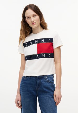 Hosszú hajú fiatal nő, aki fehér Tommy Jeans pólót visel piros és tengerészkék logóval, kék farmerral, fehér háttér előtt.