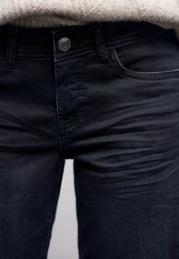 Vaqueros de mezclilla azul oscuro con textura suave, corte ajustado, diseño de cinco bolsillos y cierre con botón metálico. Desvanecimiento sutil en los muslos.