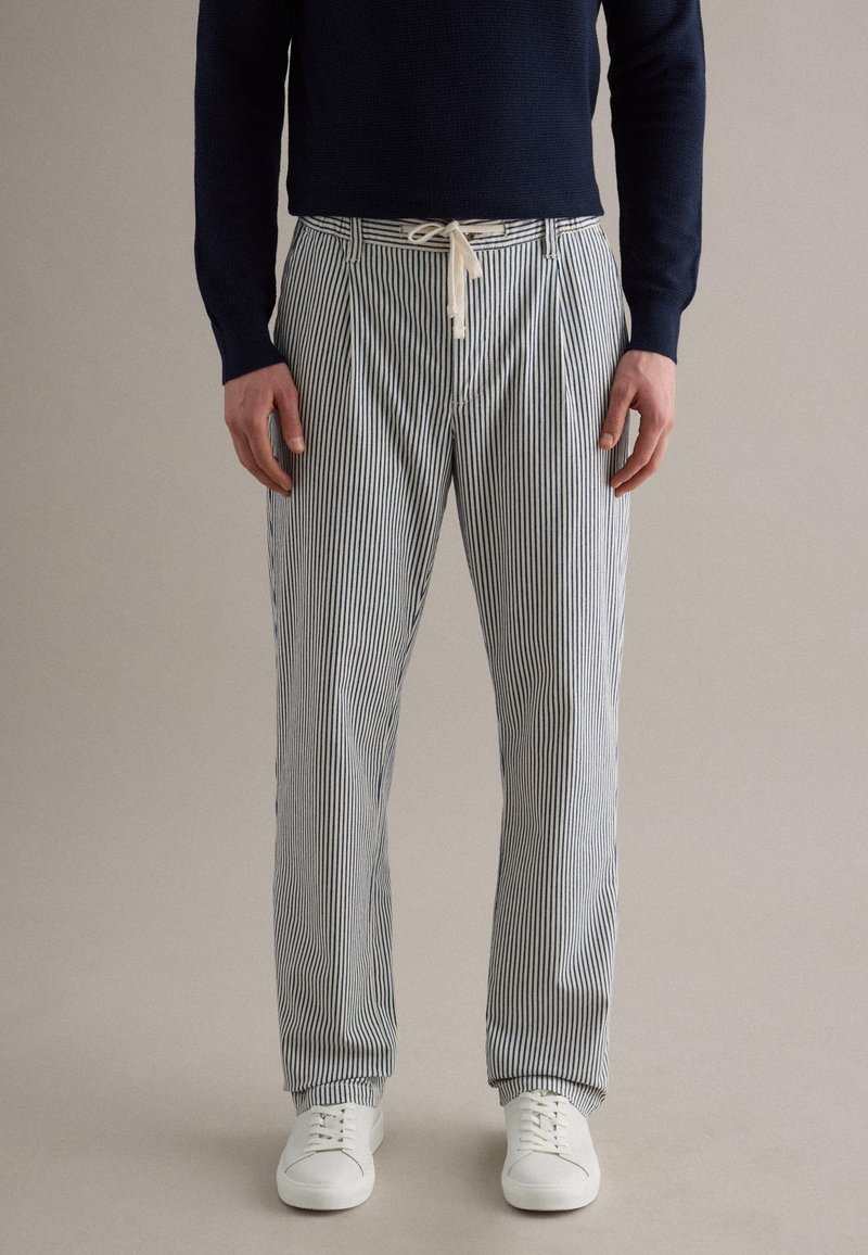 Uomo che indossa pantaloni a righe verticali bianche e blu navy con coulisse bianca, maglione blu navy e scarpe da ginnastica bianche, in piedi contro uno sfondo semplice.