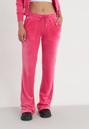 Mujer con pantalones de chándal de terciopelo rosa brillante con bolsillos delanteros y zapatillas negras, de pie con una mano en el bolsillo contra un fondo liso.