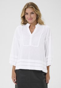 Blusa bianca con bottoni, colletto arricciato, maniche a tre quarti e texture a righe orizzontali. Presenta dettagli sovrapposti e una vestibilità ampia.