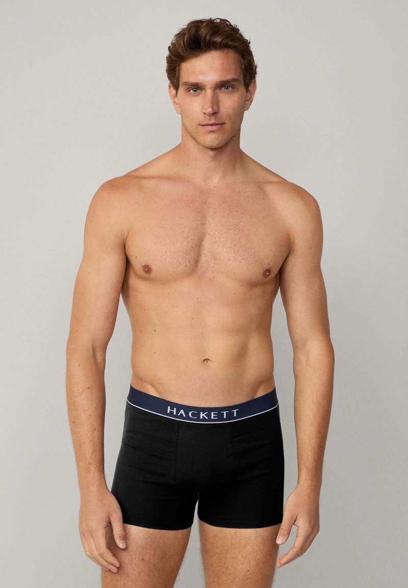 Boxer neri realizzati in tessuto morbido con un cinturino blu scuro che presenta il marchio bianco "HACKETT". La vestibilità è aderente e minimalista.