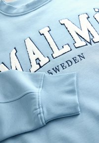 Világoskék pulóver, amelyen fehér, texturált anyagból készült "MALMÖ" felirat szerepel tengerészkék körvonallal, bordázott mandzsettákkal és gallérral, sima textúrával.
