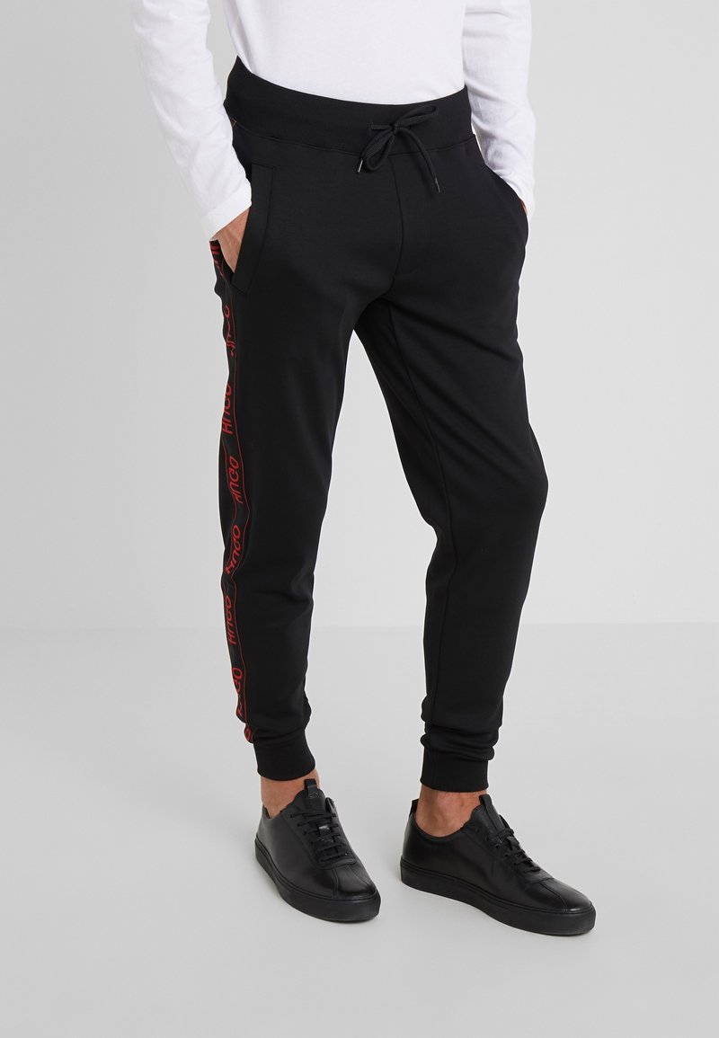 Schwarze Jogginghosen mit schmaler Passform, ausgestattet mit einem Tunnelzugbund, Seitentaschen und roten Logoakzenten entlang der Beine. Glatte Stofftextur.