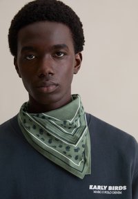 Zielony bandana z wzorem i białymi akcentami, zawiązany wokół szyi, noszony na granatowej bluzie. Bandana zdobiona jest kwiatowymi wzorami i paskami.