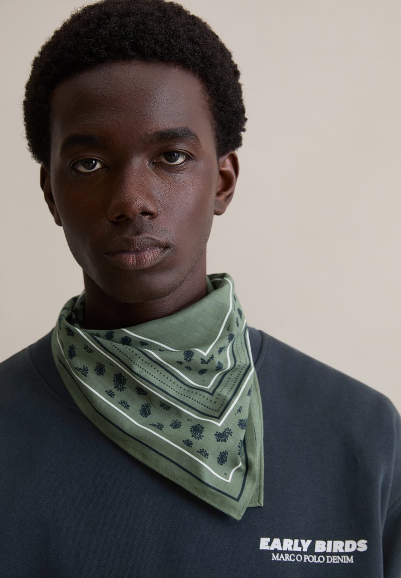 Zielony bandana z wzorem i białymi akcentami, zawiązany wokół szyi, noszony na granatowej bluzie. Bandana zdobiona jest kwiatowymi wzorami i paskami.