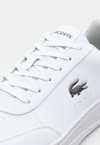 Gros plan d'une sneaker blanche Lacoste avec des lacets blancs, montrant le logo crocodile noir sur le côté et le texte Lacoste sur la languette.