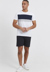 T-shirt blanche avec des rayures horizontales bleu marine et grises, associée à un short foncé et des baskets blanches. Manches courtes, design col rond.