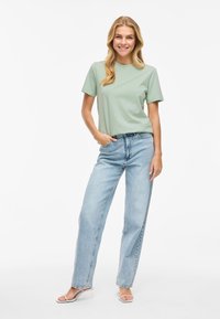 Gaiši zaļš kokvilnas T-kreklis ar īsām piedurknēm, apvienots ar brīvi pieguļošām gaiši zilām džinsu biksēm. Valkāts ar baltām šaurām sandālēm.