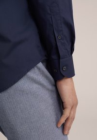 Mornarsko modra srajca z gumbi, gladke teksture in dolgimi rokavi, z okroglimi manšetami z dvema gumboma in subtilnim ukrivljenim robom.