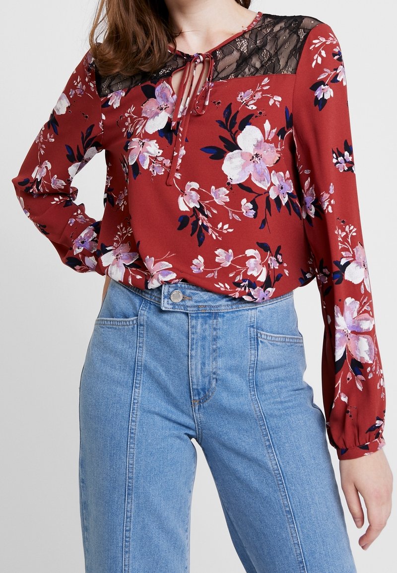 Röd blus med blommönster och spetsdetalj, rund halsringning och frontknut, som matchas med högmidjade blå denimjeans. Långa ärmar med gathered manschetter.