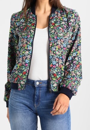 Femme portant une veste bomber fleurie colorée avec des poignets et un col noirs, par-dessus un haut blanc côtelé et un jean bleu.