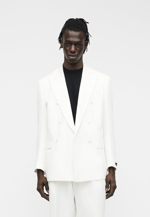 Uomo dalla pelle scura e capelli intrecciati indossa una giacca doppiopetto bianca e pantaloni bianchi sopra una camicia nera, in piedi davanti a uno sfondo semplice.