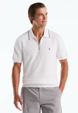 Man wearing white short-sleeve textured polo shirt with zipper and light gray pants, standing with one hand in pocket, looking right.