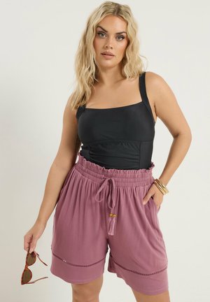 Frau mit langen blonden Haaren trägt ein schwarzes ärmelloses Oberteil und hoch taillierte mauvefarbene Shorts, hält eine Schildpatt-Sonnenbrille in einer Hand.