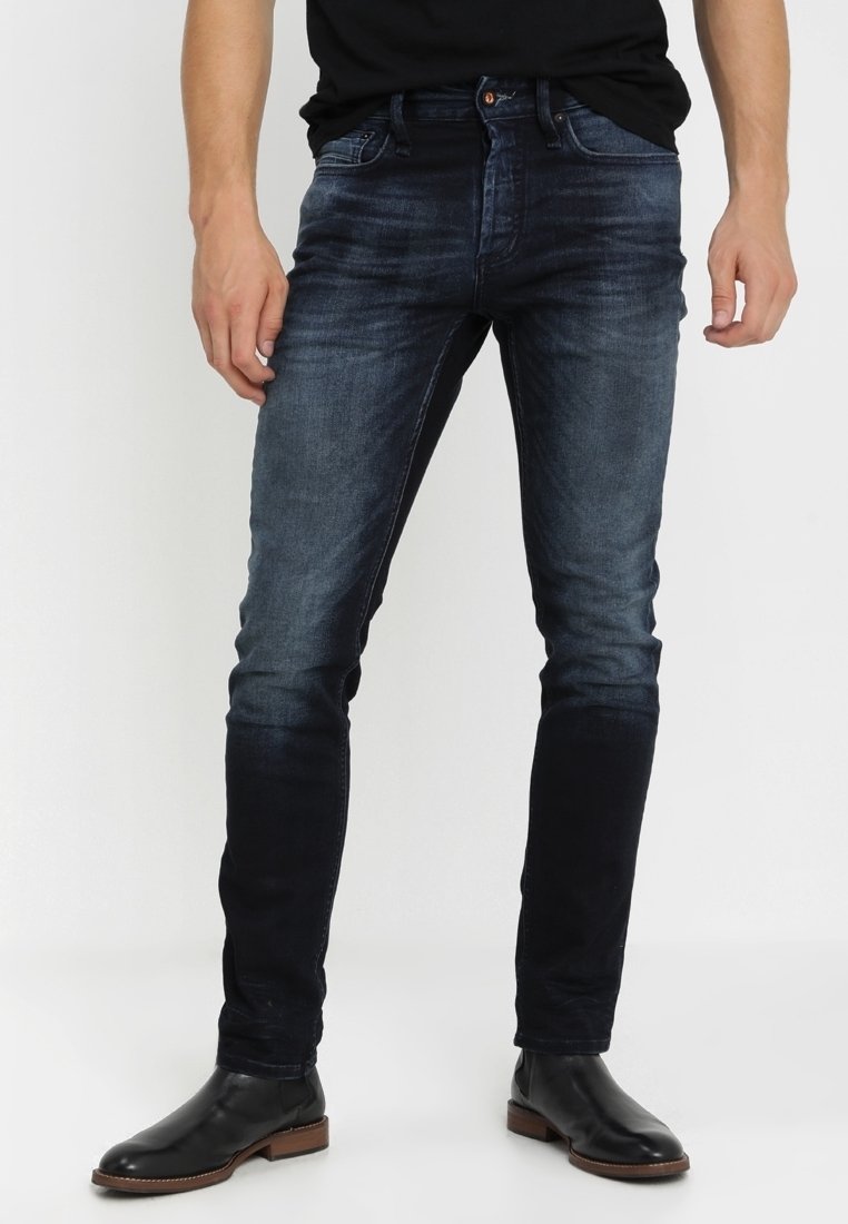 Mörkblå denimjeans med en tapered fit, som har en subtil blekning, fem fickor och en knappstängning i midjan. Bärs med svarta läderstövlar.