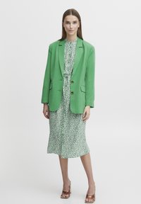 Blazer verde con dos botones marrones sobre un vestido con estampado verde y blanco. La modelo lleva tacones marrones de textura. Fondo limpio y neutral.
