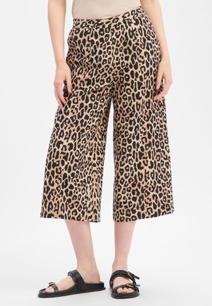 Personne portant un pantalon large beige et noir à imprimé léopard, court, avec des sandales noires à bout ouvert sur fond blanc.