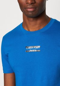 Modré bavlněné tričko s krátkými rukávy. Na střední části se nachází potištěné logo v bílé a černé barvě s nápisem "Calvin Klein Jeans" na texturovaném pozadí.